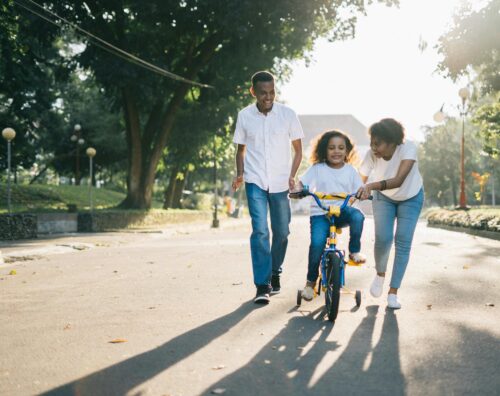 Two parents walking through the street helping their child ride a bicycle with training wheels