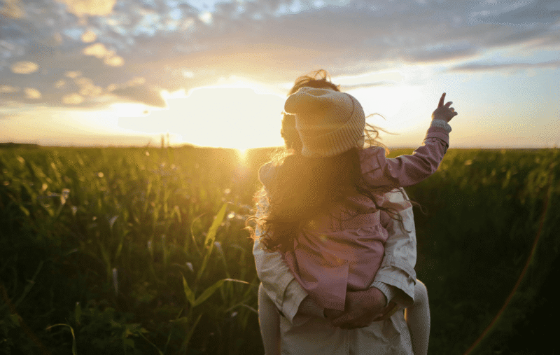a parent holds a child in a field against a sunset