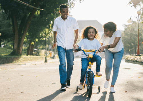 two adults coach a child on a bicycle with training wheels